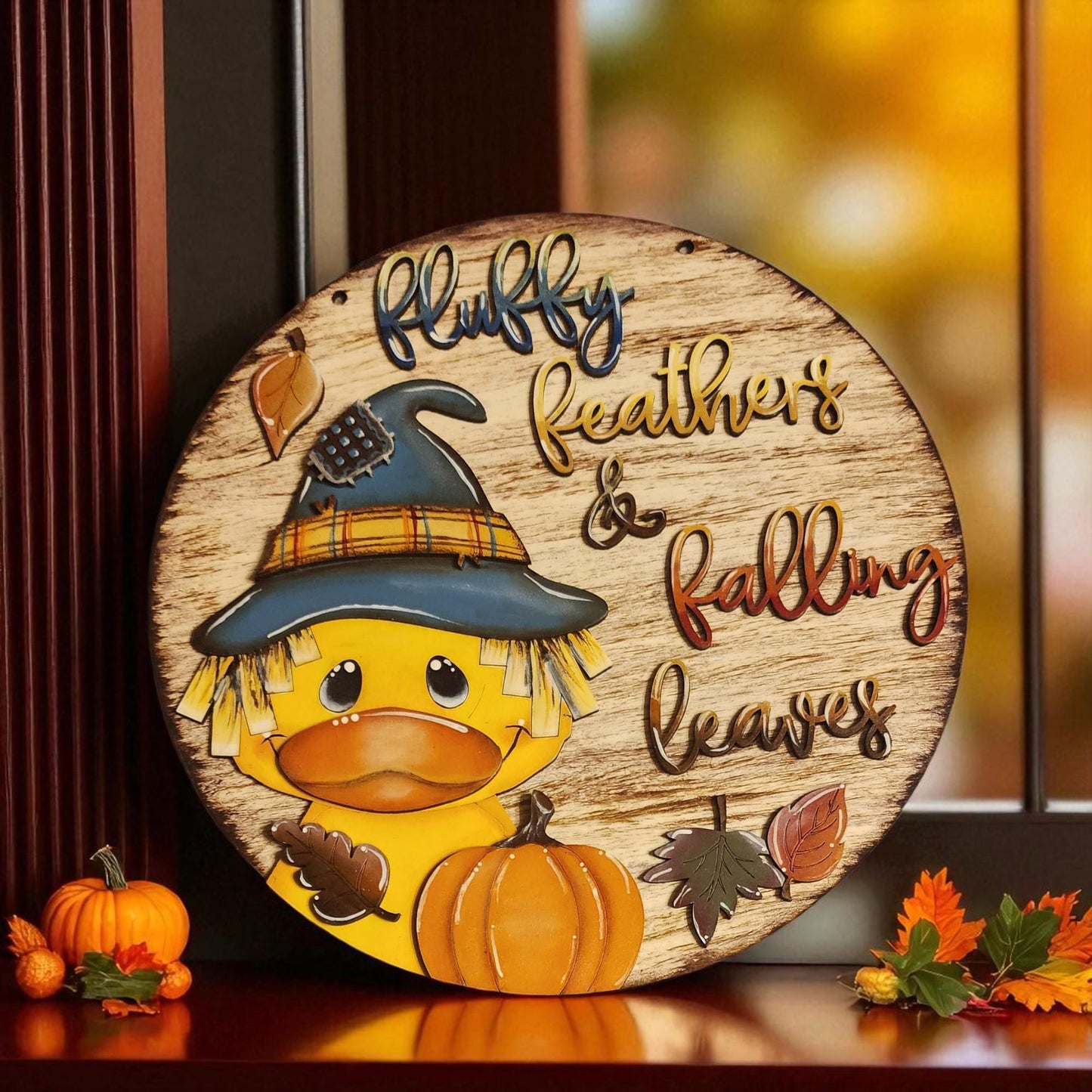 A wooden sign featuring a cartoon duck wearing a witch's hat and holding a pumpkin, with the text "Fluffy Feathers & Falling Leaves" on it. The sign is placed on a wooden surface, surrounded by autumn leaves and a pumpkin.
