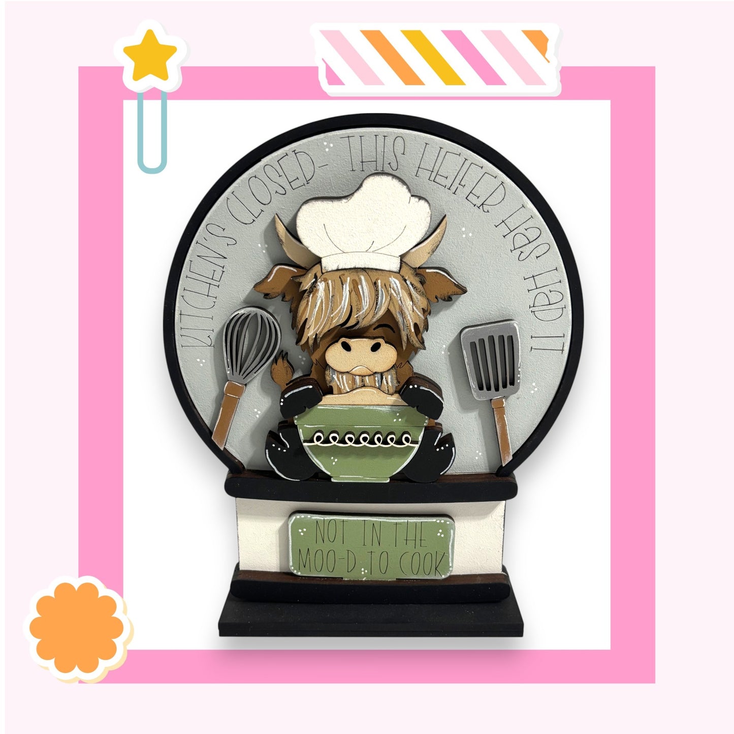 A decorative snow globe with a cartoon cow wearing a chef's hat and holding a spoon and a bowl, surrounded by kitchen utensils.