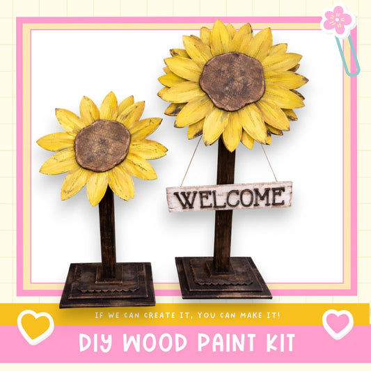 two wooden sunflower sculptures, each with a wooden sign attached to them. The sign says "WELCOME". The sculptures are placed on a wooden stand, and there is a DIY wood paint kit visible in the image.