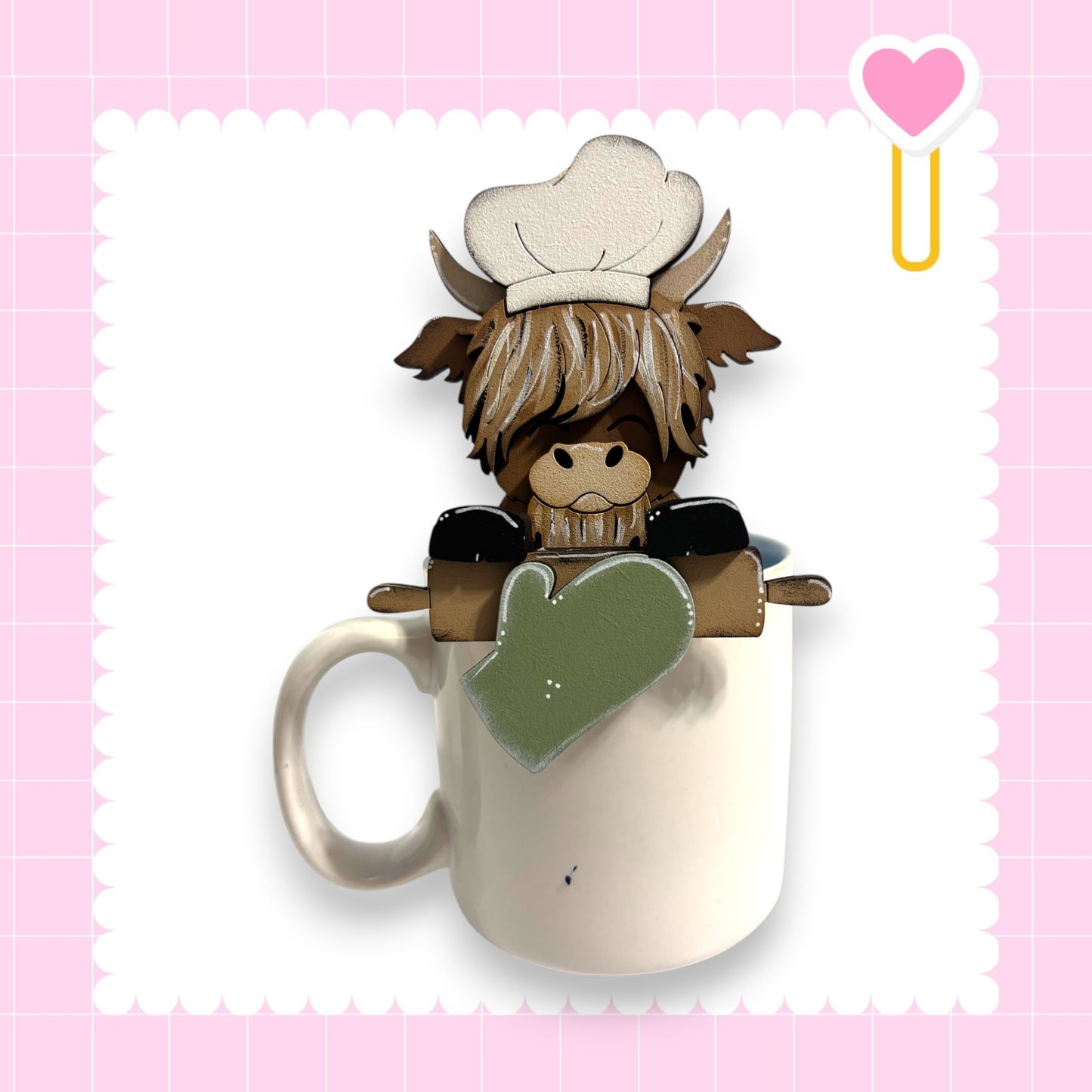 A cartoon cow wearing a chef's hat and holding a rolling pin is sitting inside a white mug, set against a pink background with a heart shape.