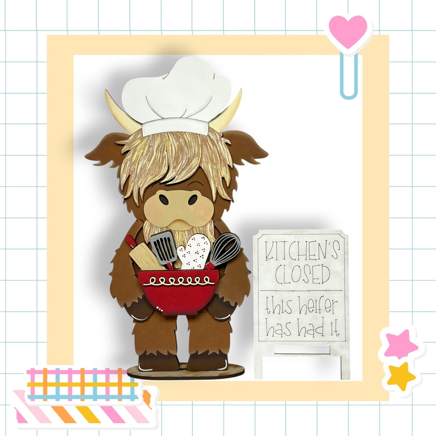 A wooden cutout of a cow wearing a chef's hat and holding a red bowl with kitchen utensils inside. The cow is standing in front of a sign that says "Kitchen's Closed".