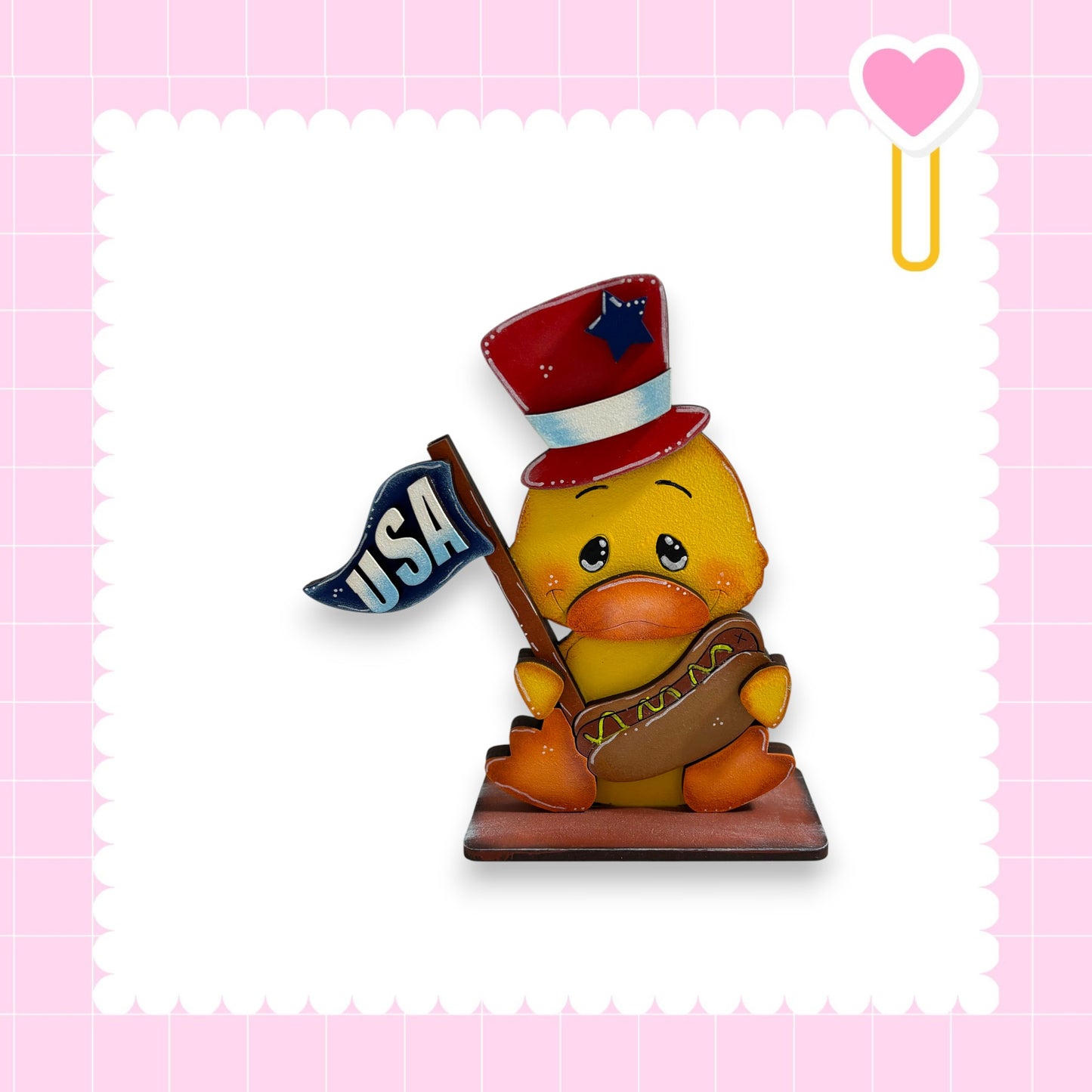 A yellow duck wearing a red hat with a blue star and holding a blue flag with the word "USA" on it, sitting on a brown base.