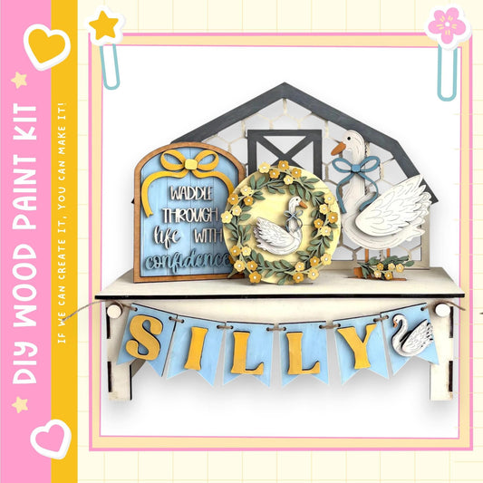 The image is a wooden paint kit featuring a decorative wooden sign with a wreath, a wooden sign with the word "Silly" written on it, and a wooden sign with a goose. The kit appears to be a creative and playful display, possibly for a child's room or a nursery.