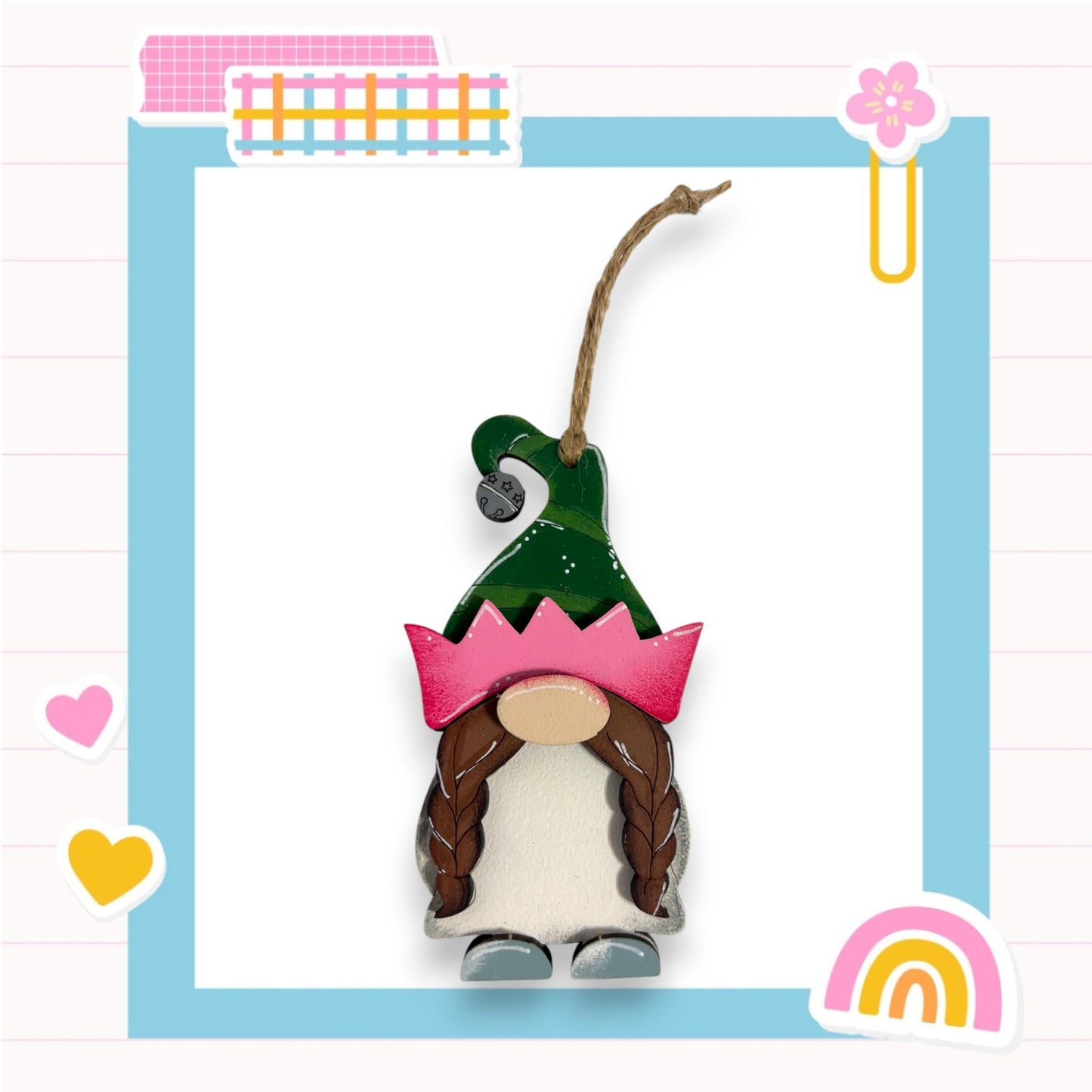 A small wooden gnome figure wearing a green hat with a pink crown, set against a blue background with various decorative elements such as hearts, a rainbow, and a flower.