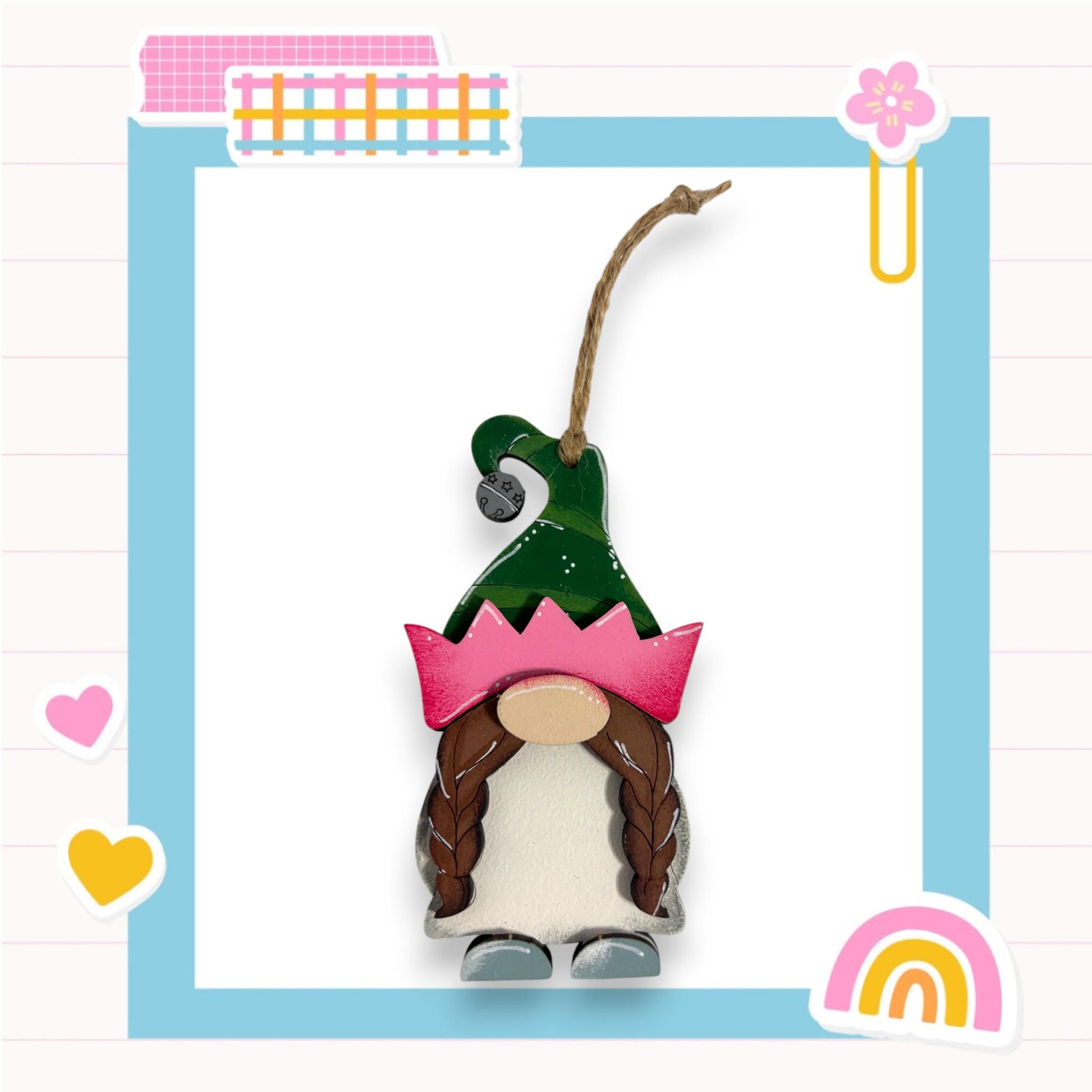 A small wooden gnome figure wearing a green hat with a pink crown, set against a blue background with various decorative elements such as hearts, a rainbow, and a flower.