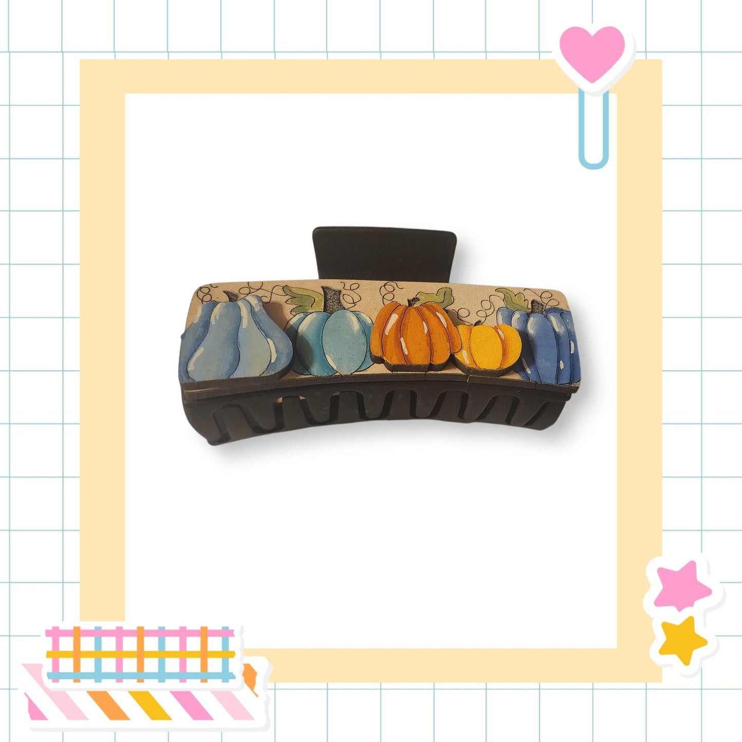 a black hair clip with a pumpkin and blue squash design on it, set against a white background with a grid pattern.
