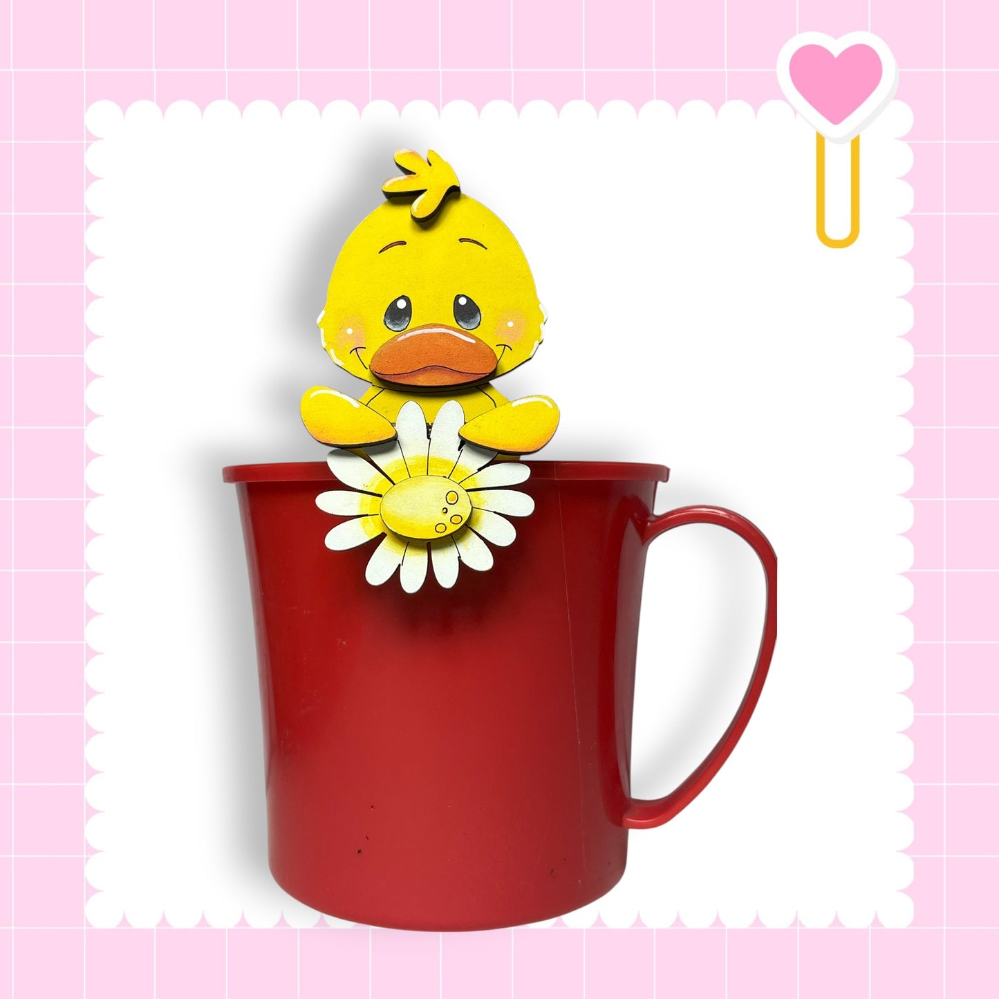 A red mug with a yellow duck figurine holding a flower on top, set against a pink background with a heart shape.