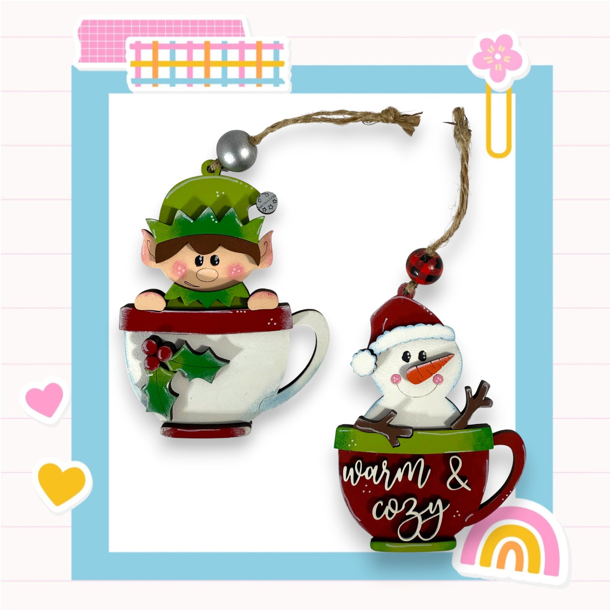 two Christmas ornaments, one of a elf and one of a snowman, both sitting inside a cup. The elf is wearing a green hat and the snowman is wearing a red hat. The cup has the words "Warm & Cozy" written on it. The background of the image is a light blue color with a pink and white checkered pattern.