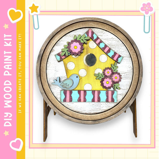 a wooden birdhouse with a yellow body, pink and blue accents, and a blue bird perched on the roof. The birdhouse is displayed on a stand, and there are flowers and leaves surrounding it. The image also includes a heart and a star, adding a decorative touch.