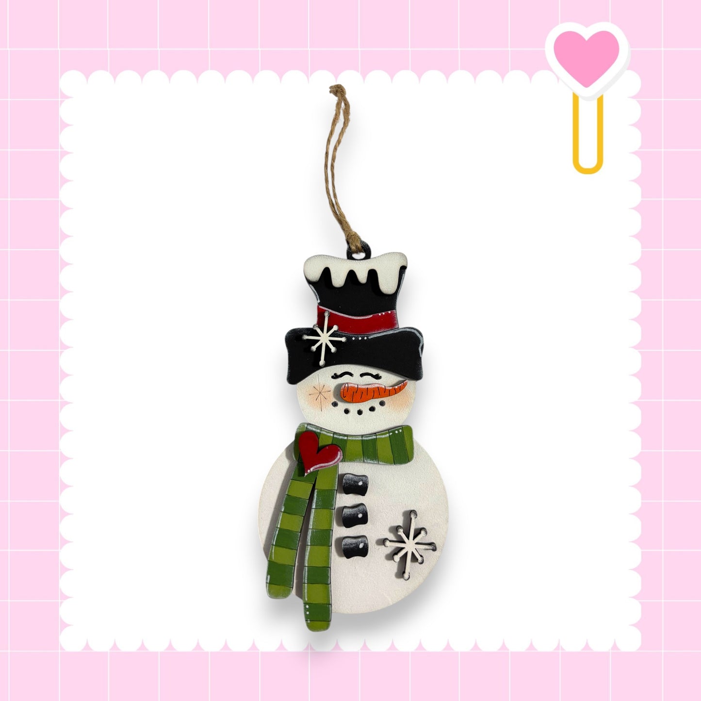 A snowman ornament with a black top hat, green scarf, and red scarf is hanging against a pink background with a scalloped edge.