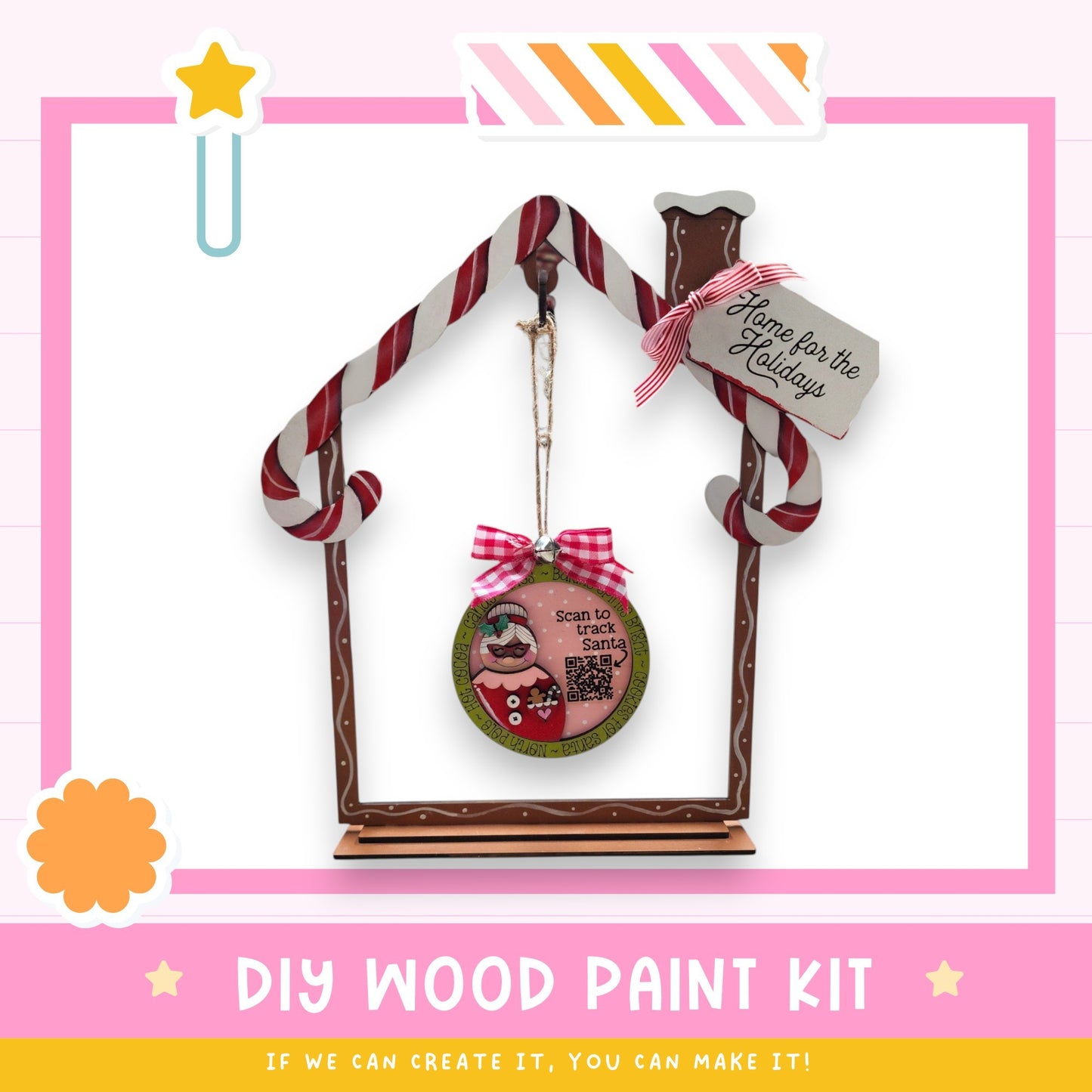 a DIY wood paint kit for creating a wooden ornament in the shape of a house. The kit includes a wooden frame, a candy cane decoration, and a sign that says "Home for the Holidays". The kit is presented in a pink and white color scheme, with a star decoration on the left side. The text "DIY WOOD PAINT KIT" is displayed at the bottom of the image, along with the phrase "If we can create it, you can make it!"