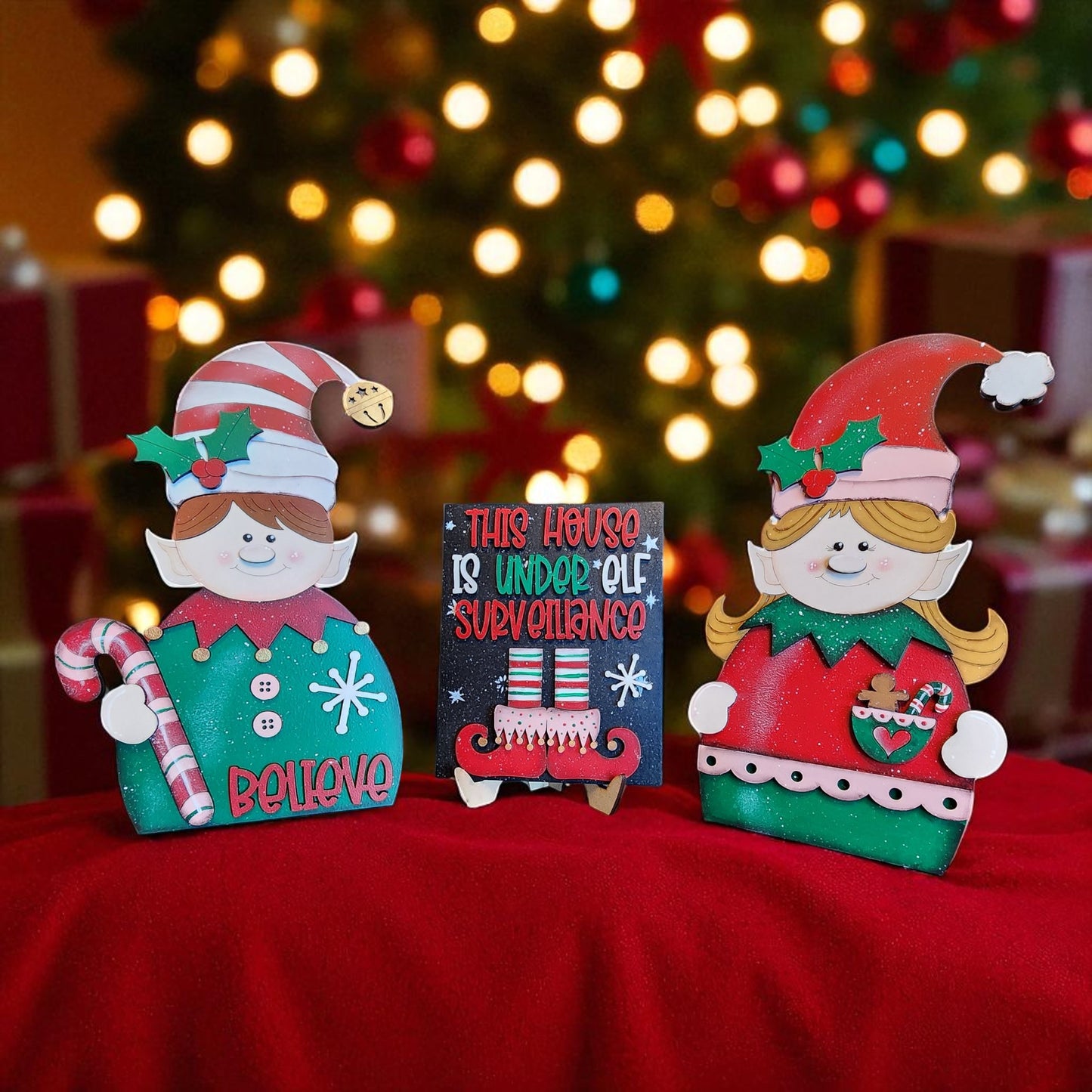 two wooden elf figurines and a sign with text, all placed on a red cloth against a blurred background with Christmas lights.