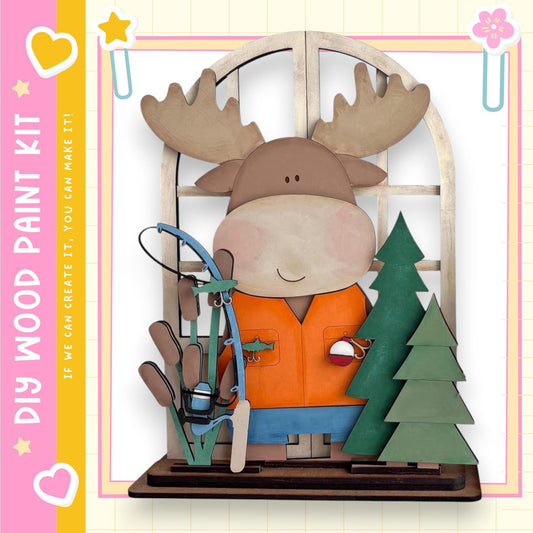 A wooden toy moose holding a fishing rod and standing next to a Christmas tree, with a pink and yellow border around the image.