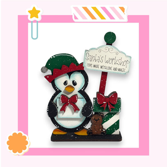 A wooden toy penguin wearing a Santa hat and holding a gift box, standing next to a sign that says "Santa's Workshop Toys Made with Love and Magic".