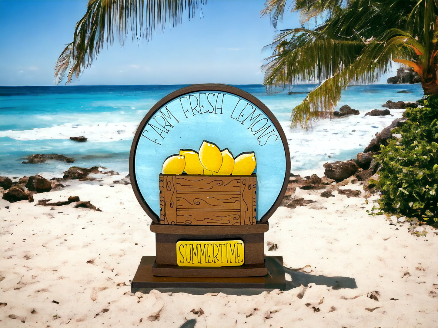 A circular sign on a sandy beach with a blue background, featuring a wooden crate with lemons and the text "Farm Fresh Lemons Summertime"