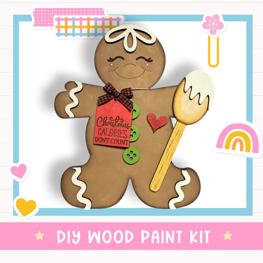 A wooden gingerbread man figure with a red tag that says "Christmas Calories Don't Count" and a heart-shaped button on its chest. The gingerbread man is holding a spoon and is surrounded by various colorful stickers and a rainbow. The image is described as a DIY wood paint kit.