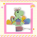 A green dinosaur toy is sitting on top of a pile of colorful Easter eggs, with a tag attached to the dinosaur that reads "Easter dinosaur Rex".