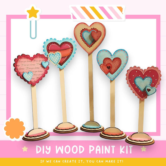 The image is a DIY wood paint kit featuring a collection of heart-shaped wooden sticks arranged in a row, with a pink and white background.