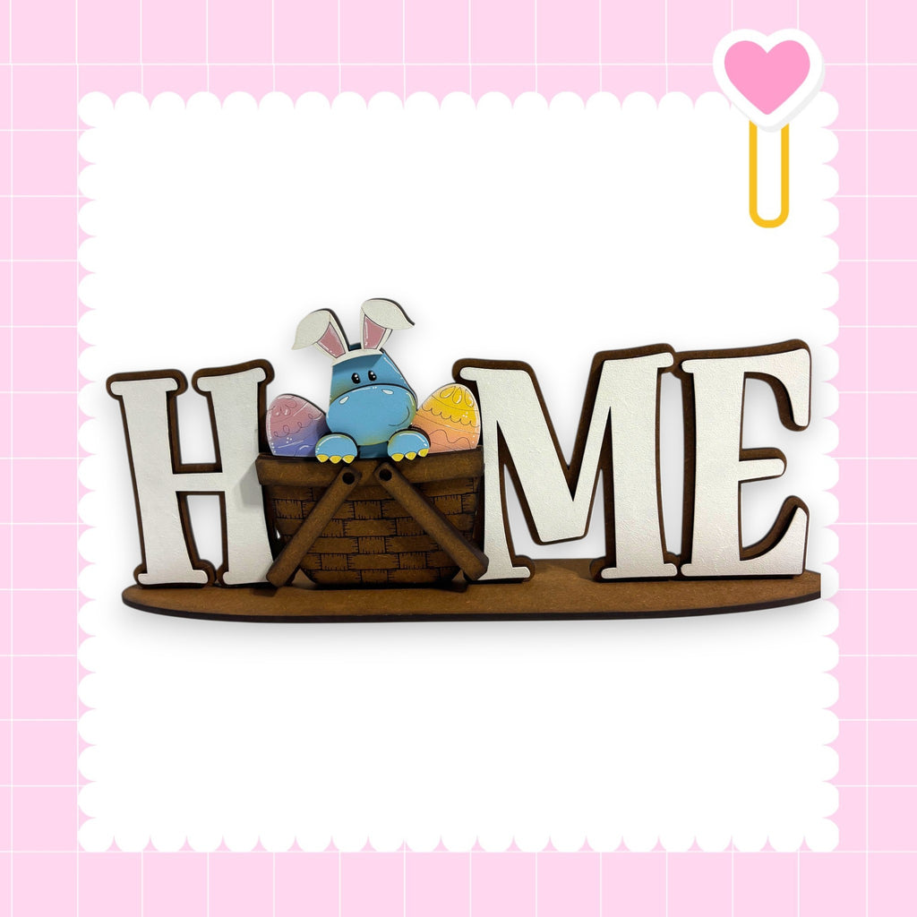 A wooden sign with the word "HOME" in large letters, featuring a small blue bunny sitting in a basket.