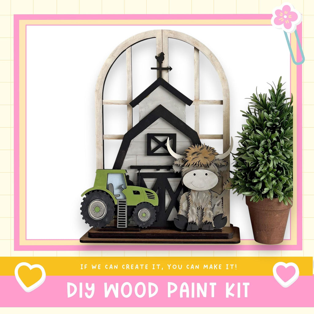 a DIY wood paint kit, including a wooden barn, tractor, and a stuffed animal cow. The barn is made of wood and has a black roof, while the tractor is painted green. The cow is also painted green and has a brown mane. The image is framed by a pink border, and there is a heart and a flower in the corners. The text at the bottom of the image reads "If we can create it, you can make it!"