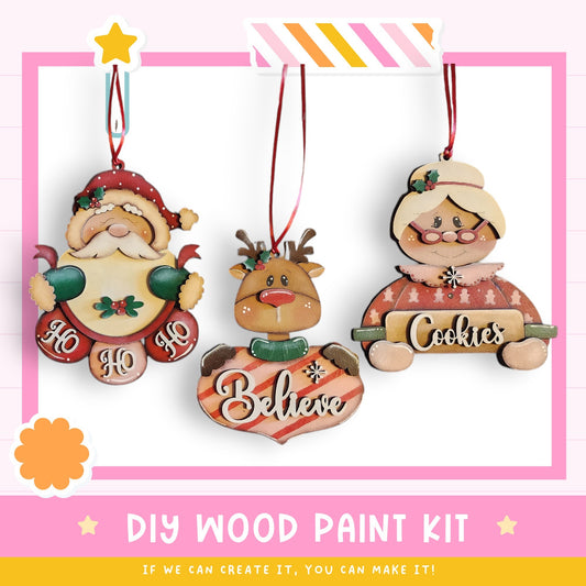 three wooden ornaments hanging on a pink background. The ornaments include a Santa Claus figure, a reindeer, and a woman holding a sign that says "Cookies". The image also includes text that says "DIY WOOD PAINT KIT" and "IF WE CAN CREATE IT, YOU CAN MAKE IT", suggesting that the ornaments can be created with a paint kit.