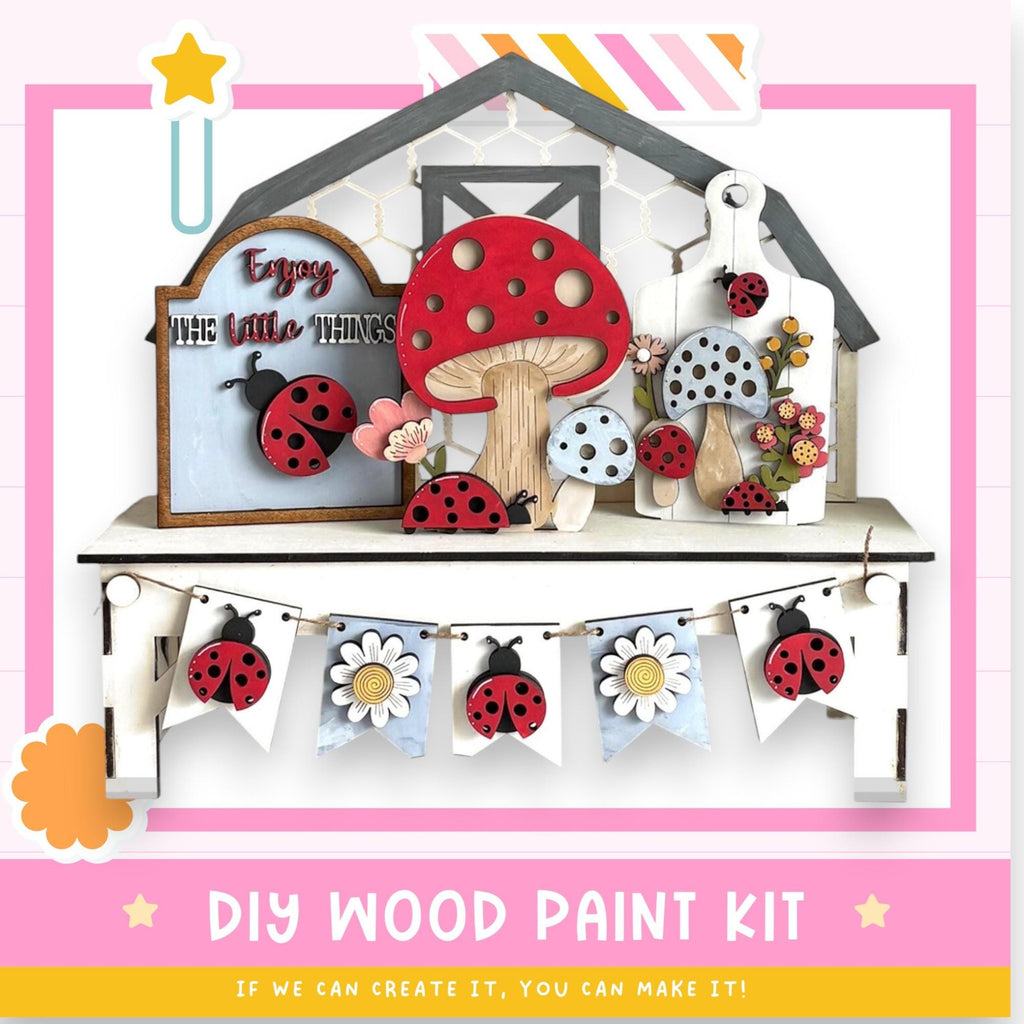 The image is a colorful and whimsical DIY wood paint kit featuring a wooden house, mushrooms, ladybugs, and a banner with flowers. The kit appears to be designed for children to create their own wooden decorations.