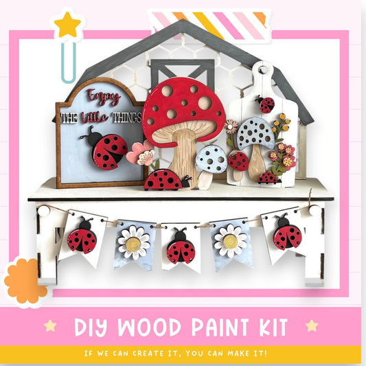 The image is a colorful and whimsical DIY wood paint kit featuring a wooden house, mushrooms, ladybugs, and a banner with flowers. The kit appears to be designed for children to create their own wooden decorations.