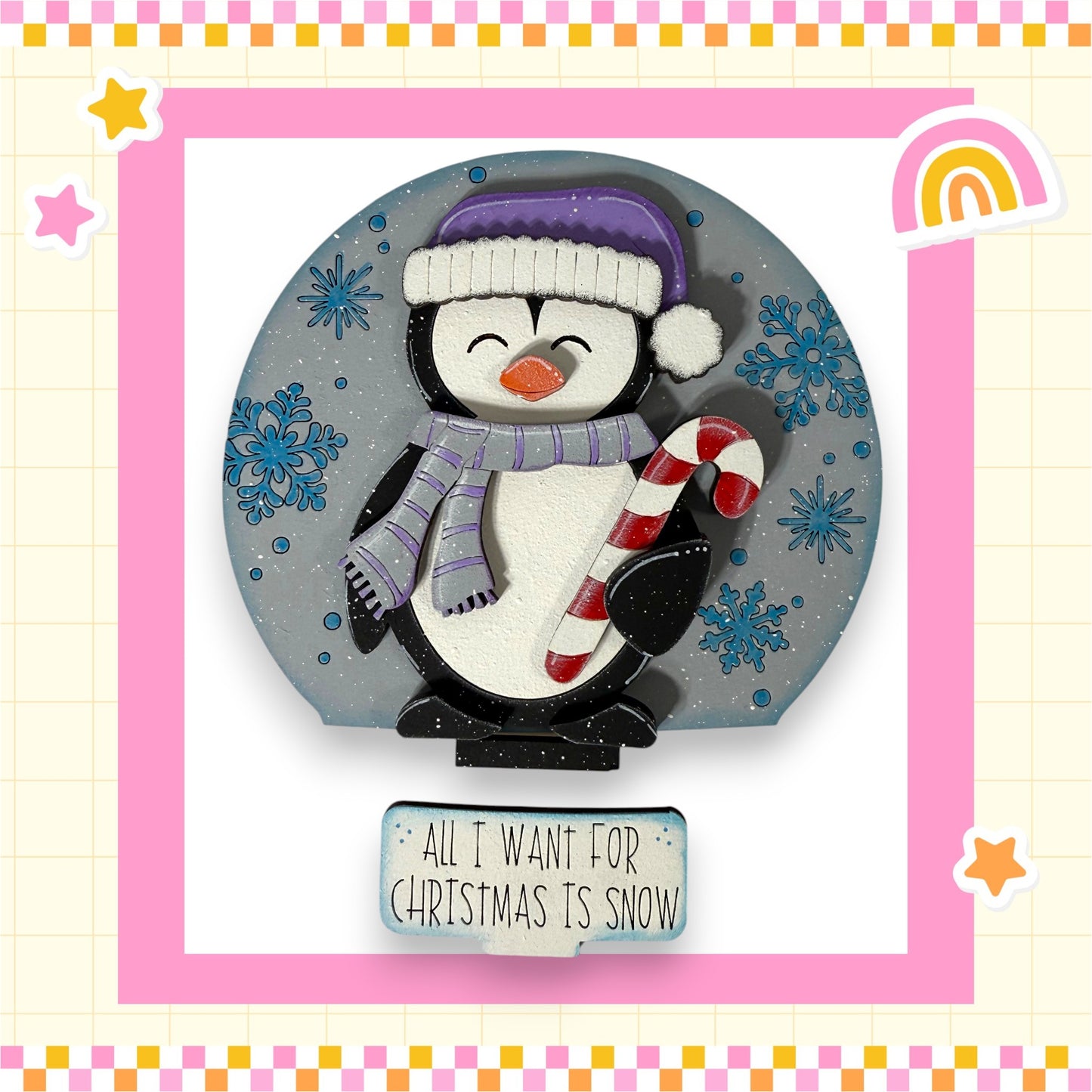 A penguin wearing a purple hat and scarf, holding a red and white striped candy cane, is surrounded by snowflakes and a rainbow, with a sign that says "All I want for Christmas is snow".