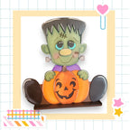 A carved pumpkin with a carved face and a green monster figure holding it, set against a grid background with a heart and a star.