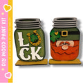 two mason jars with painted designs on them, including a green jar with the word "LUCK" and a red jar with a smiling face and a clover.