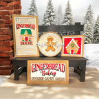 a wooden bench with three framed signs advertising a gingerbread house bakery. The signs display the bakery's name, "Gingerbread Bakery", and its offerings, "Cookies & Candy Gakes". The signs are set against a snowy background with pine trees, creating a festive and cozy atmosphere.