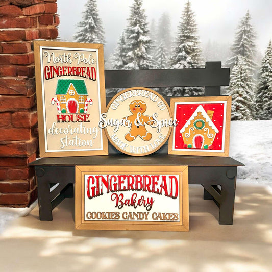 a wooden bench with three framed signs advertising a gingerbread house bakery. The signs display the bakery's name, "Gingerbread Bakery", and its offerings, "Cookies & Candy Gakes". The signs are set against a snowy background with pine trees, creating a festive and cozy atmosphere.
