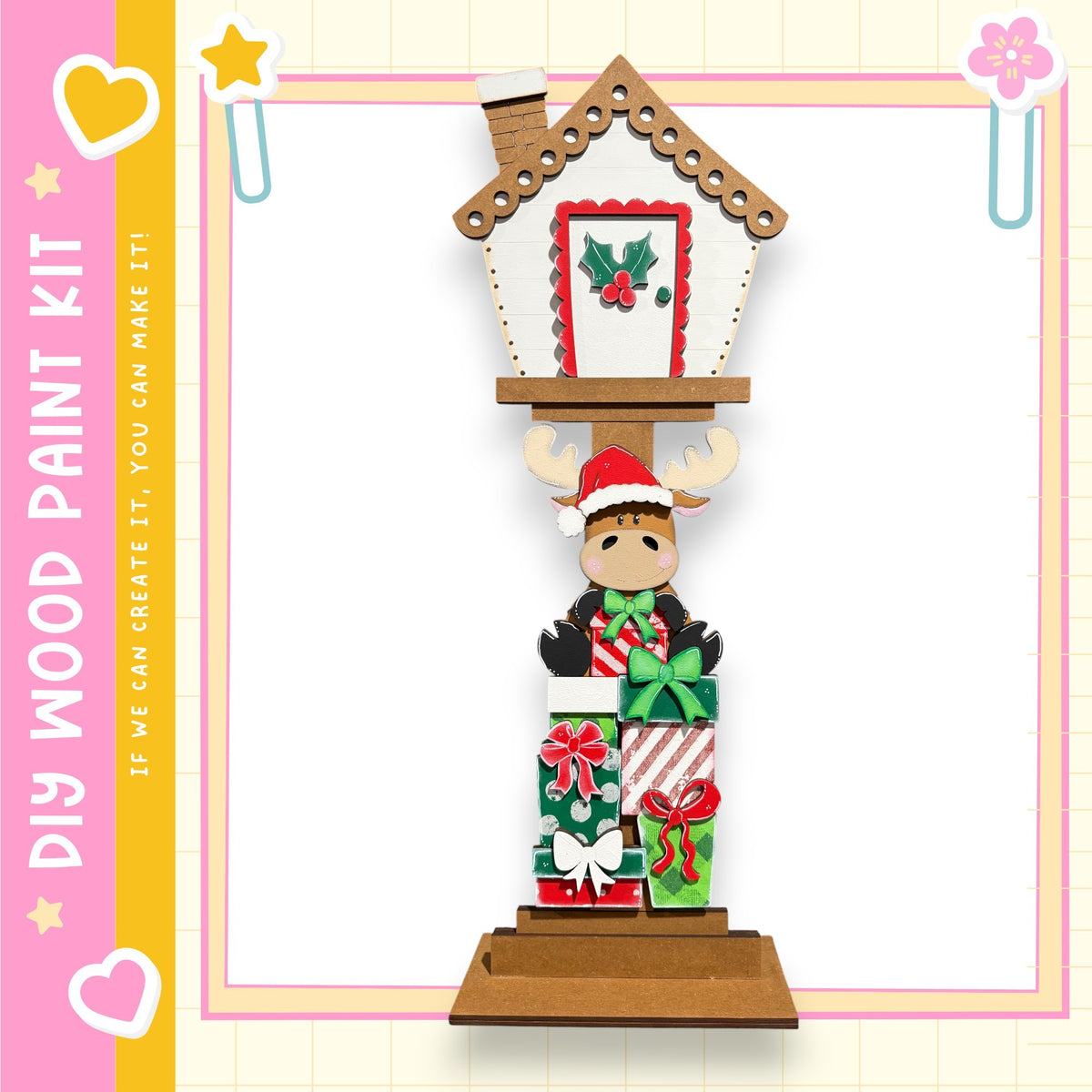 A wooden Christmas ornament with a moose figure and presents, set against a pink and yellow background with a heart and star design.