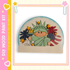 A wooden paint kit depicting the Statue of Liberty surrounded by various patriotic symbols, including a flag, fireworks, and a heart.