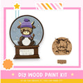 a DIY wood paint kit for creating a snow globe, with a wooden snow globe containing a cartoon cow wearing a purple hat and a sign that says "Winter is better on the farm".