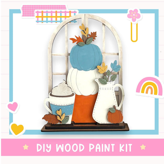 The image is a DIY wood paint kit featuring a wooden shelf with a vase, a pumpkin, and a cup of whipped cream, surrounded by various colorful stickers and a rainbow.