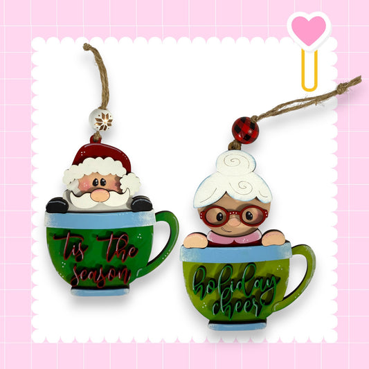 two Christmas ornaments, each shaped like a cup. One ornament is designed to resemble Santa Claus, while the other is shaped like an elderly woman. The ornaments are hanging from strings and are decorated with the text "Tis the season" and "holly cheer" written on them.