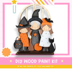 The image is a DIY wood paint kit featuring three wooden figures dressed in Halloween costumes, including a witch, a girl, and a cat, set against a pink and white background.