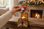 A hand is placing a candle in a gingerbread man decoration, which is sitting on a wooden surface in front of a fireplace.