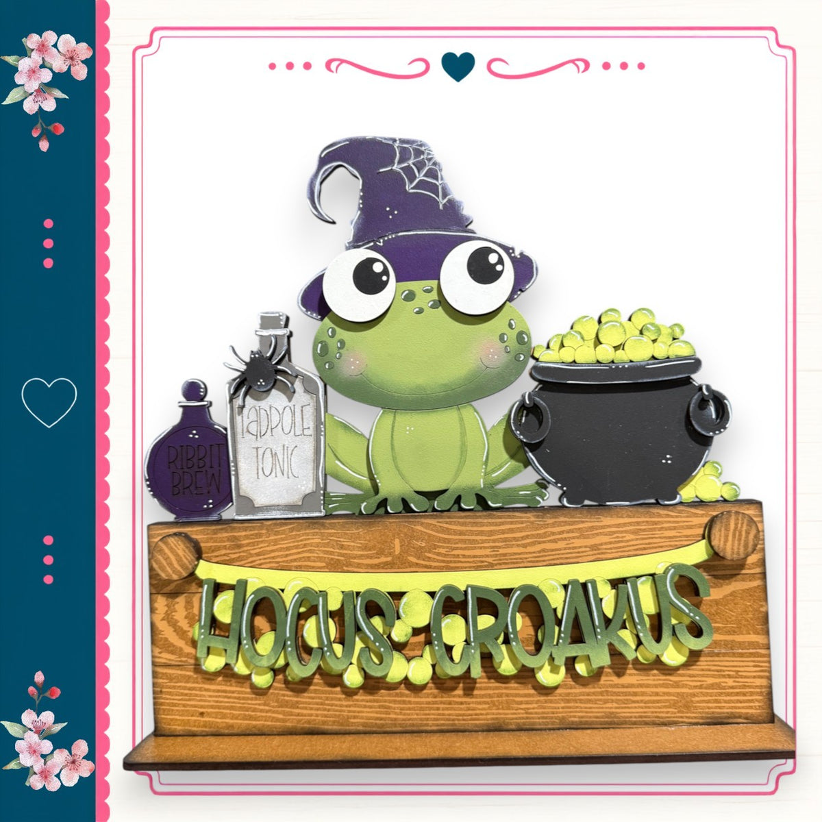 A green frog wearing a witch's hat and holding a cauldron filled with green objects, standing on a wooden shelf with the words "Hocus-Gringus" written on it.