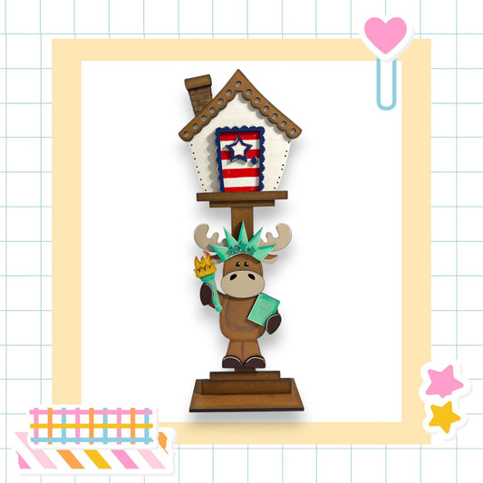 A cartoon moose holding a book stands in front of a miniature American flag-themed house, surrounded by colorful stickers and a heart-shaped object.