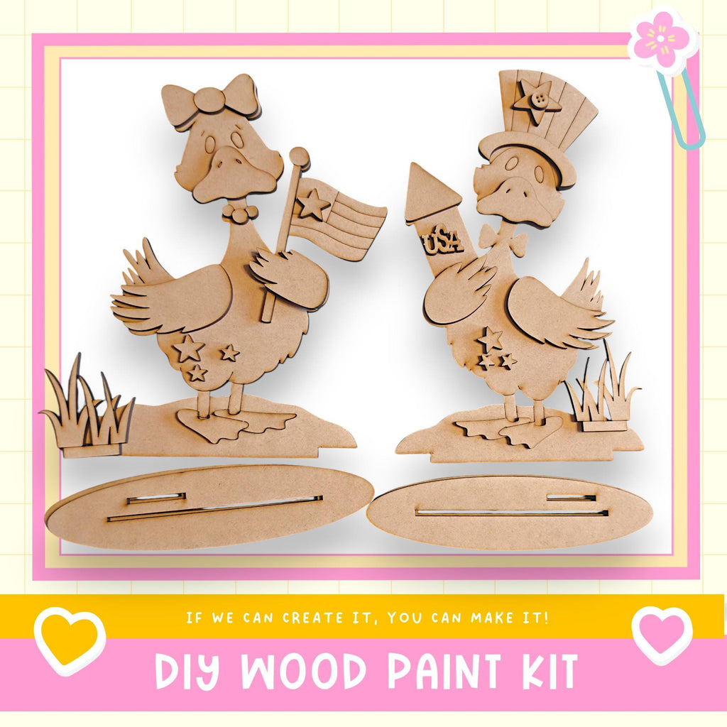 two wooden duck figures, one holding a flag and the other a rocket, standing on a platform. The text below the image reads "DIY WOOD PAINT KIT" and "If we can create it, you can make it!"