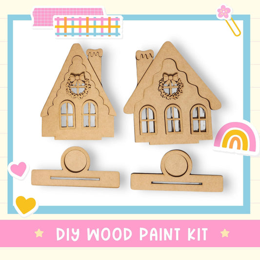 a DIY wood paint kit, including two wooden house-shaped cutouts and a heart-shaped cutout, all set against a light blue background with a pink border.
