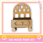 a DIY wood paint kit, with a wooden cabinet or shelf depicted in the center. The kit appears to be made of wood and has a drawer. The image is framed with a pink border and includes a heart symbol on the left side. The text "DIY WOOD PAINT KIT" is displayed at the bottom of the image.