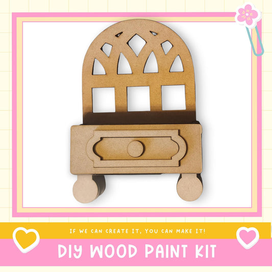a DIY wood paint kit, with a wooden cabinet or shelf depicted in the center. The kit appears to be made of wood and has a drawer. The image is framed with a pink border and includes a heart symbol on the left side. The text "DIY WOOD PAINT KIT" is displayed at the bottom of the image.