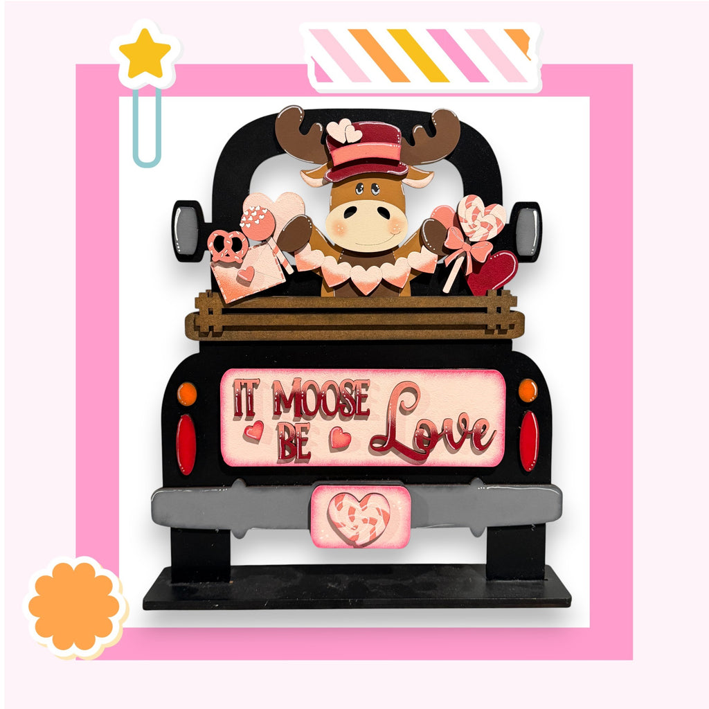 A wooden truck with a moose figure and various heart-shaped objects, including a pink heart and a red heart, on the back. The truck has a sign that says "It Moose Be Love".