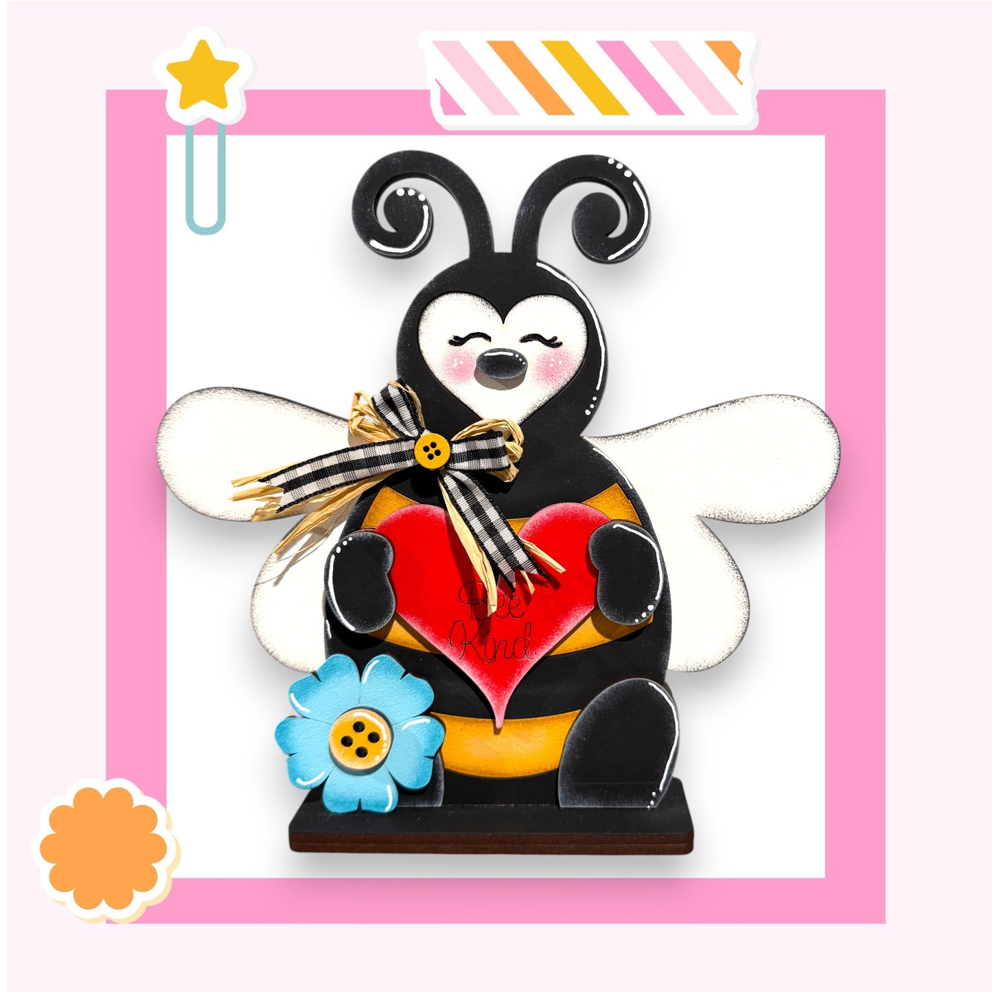 A wooden bee figurine holding a heart and a flower, with a star decoration above it, is set against a pink background.