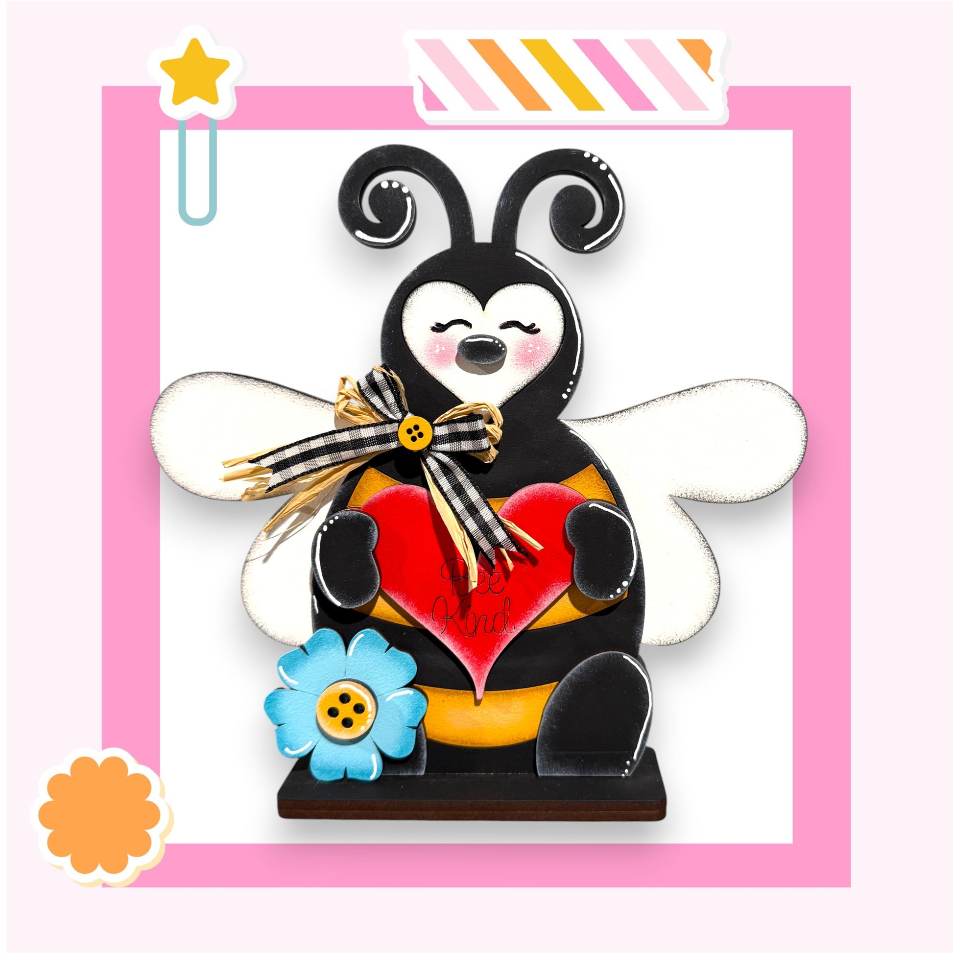 A wooden bee figurine holding a heart and a flower, with a star decoration above it, is set against a pink background.