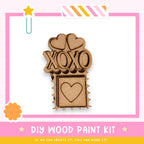a DIY wood paint kit with a heart-shaped cutout and the word "XOXO" written on it, set against a pink and white background.