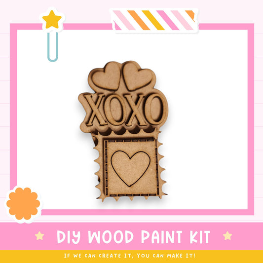 a DIY wood paint kit with a heart-shaped cutout and the word "XOXO" written on it, set against a pink and white background.